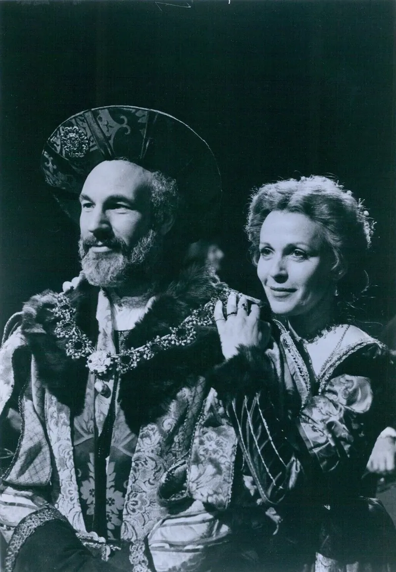 Black-and-white still of Patrick Stewart as King Claudius in a fur-trimmed royal costume and large hat, with a woman in period dress resting her hand on his shoulder